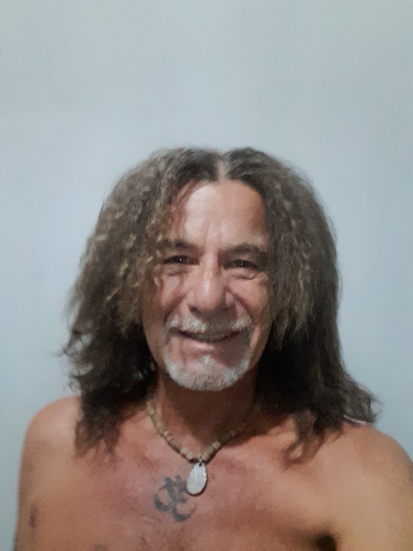 Nahaufnahme eines Mannes mit langen lockigen Haaren, grauem Bart und Halskette vor hellem Hintergrund.
